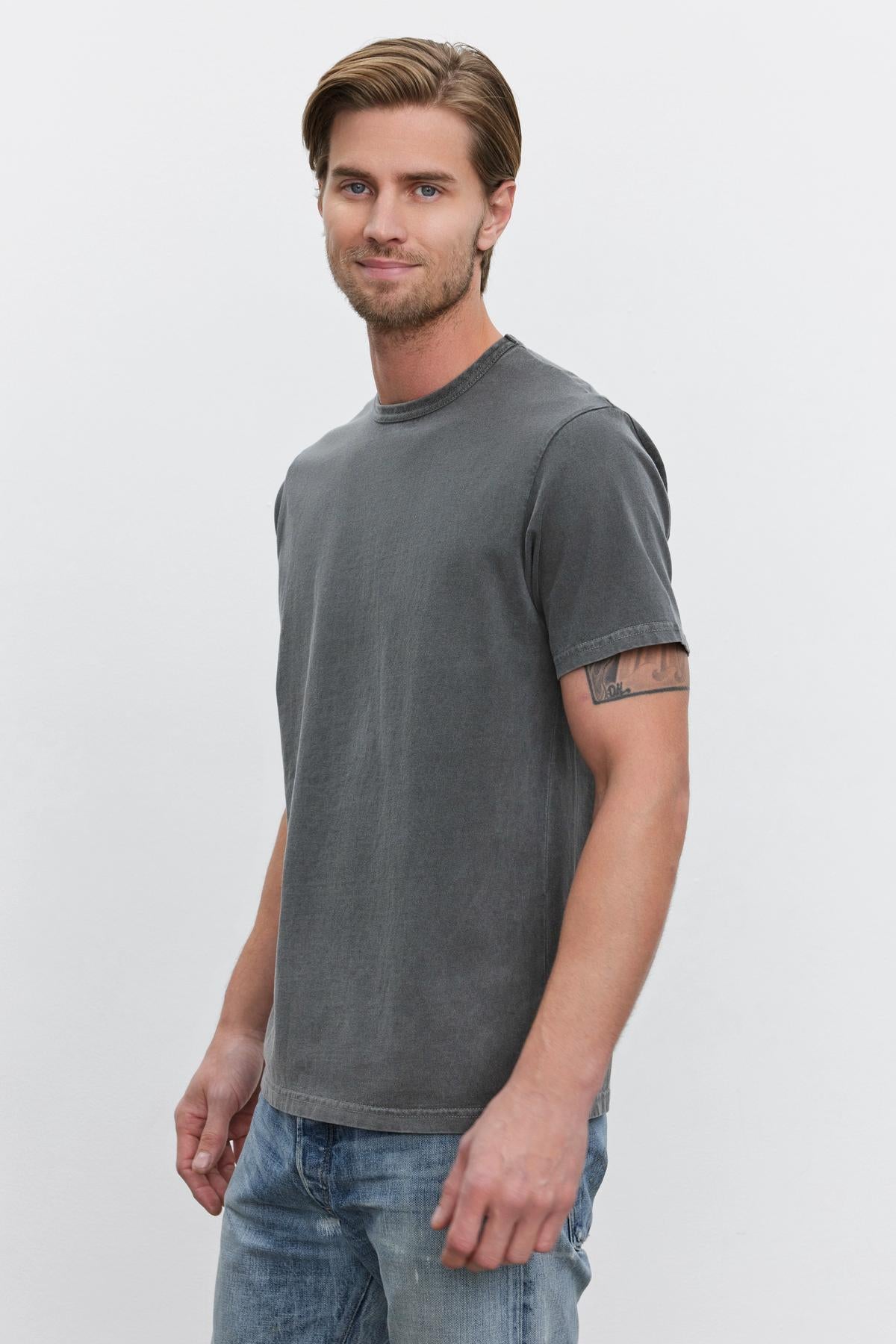   A man with light brown hair and a beard wears the STITCH TEE by Velvet by Graham & Spencer along with blue jeans. He stands in front of a plain white background and looks at the camera. 