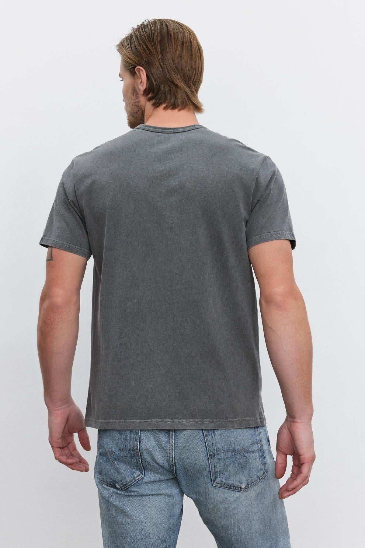   A man is standing with his back facing the camera, wearing a plain gray STITCH TEE by Velvet by Graham & Spencer and blue jeans. 