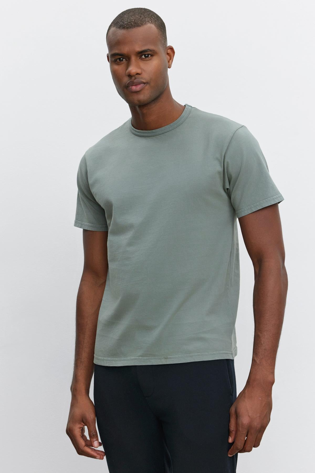   A man wearing a short-sleeved, gray-green STITCH TEE by Velvet by Graham & Spencer and black pants stands against a plain white background. 