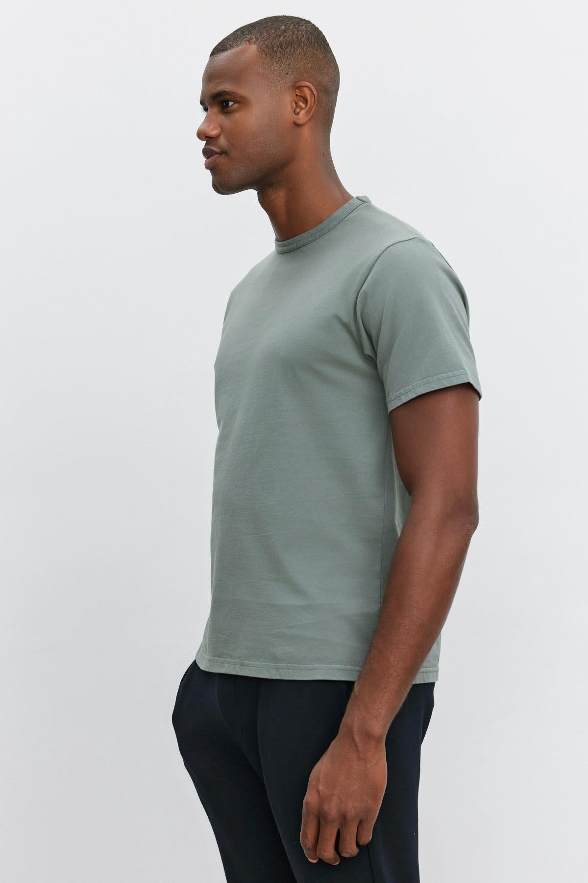   A man in a green Velvet by Graham & Spencer STITCH TEE and dark pants stands sideways against a plain white background. 