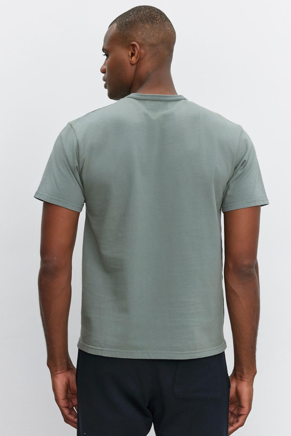   A man wearing a STITCH TEE from Velvet by Graham & Spencer and black pants stands with his back facing the camera against a plain white background. 