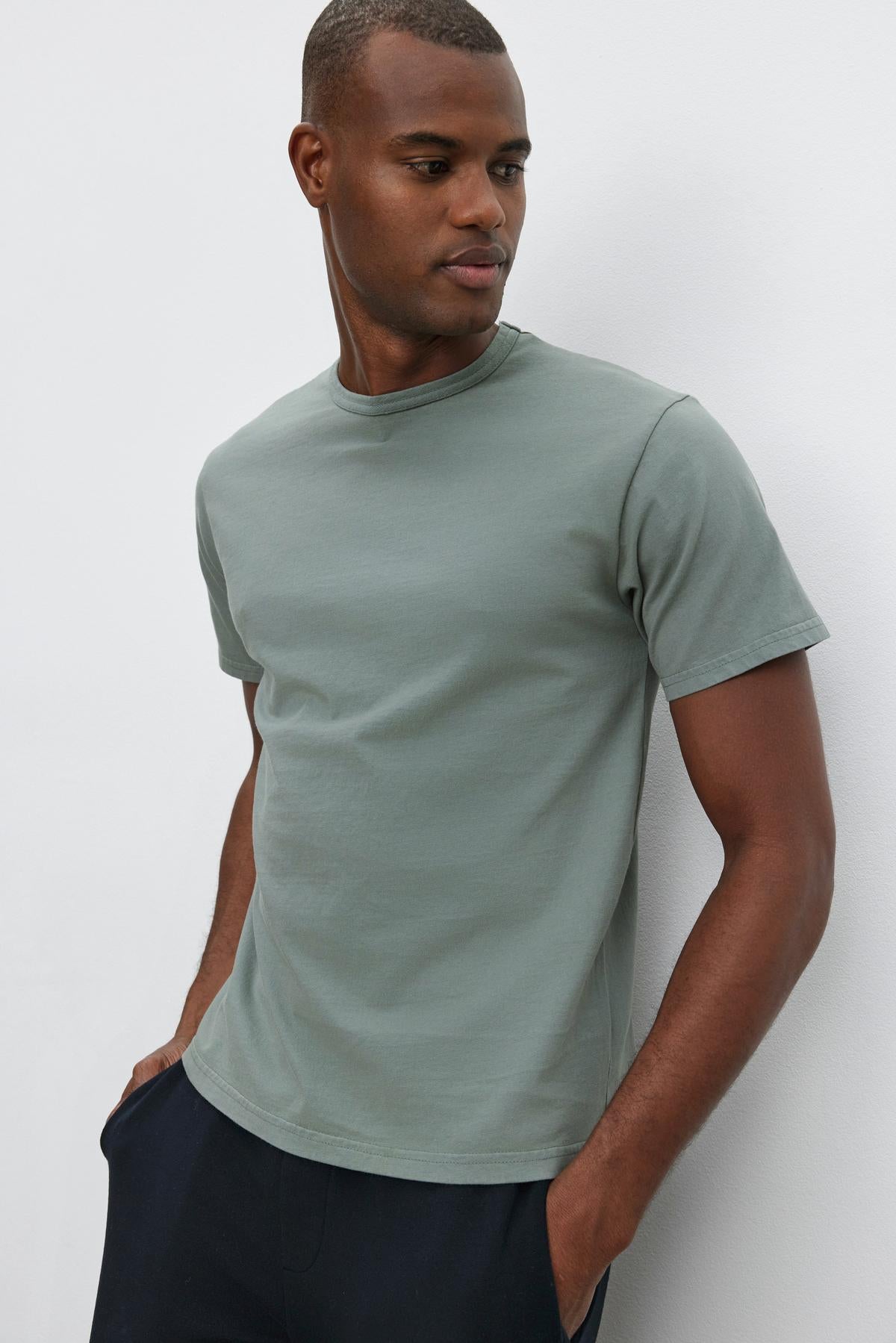   A man in a light green Velvet by Graham & Spencer STITCH TEE and dark pants stands against a plain white background, looking to his left with his hands in his pockets. 