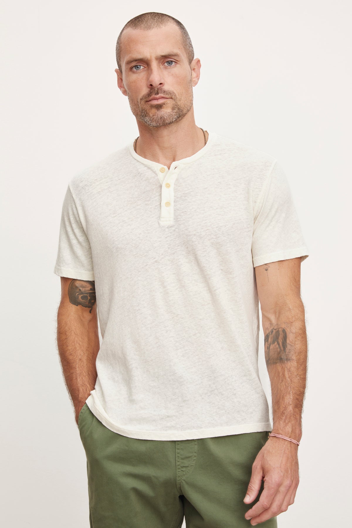 A man with a short beard and tattoos on his arms wearing a Velvet by Graham & Spencer Lionel Henley style shirt and olive green pants, standing against a white background.-36753588682945