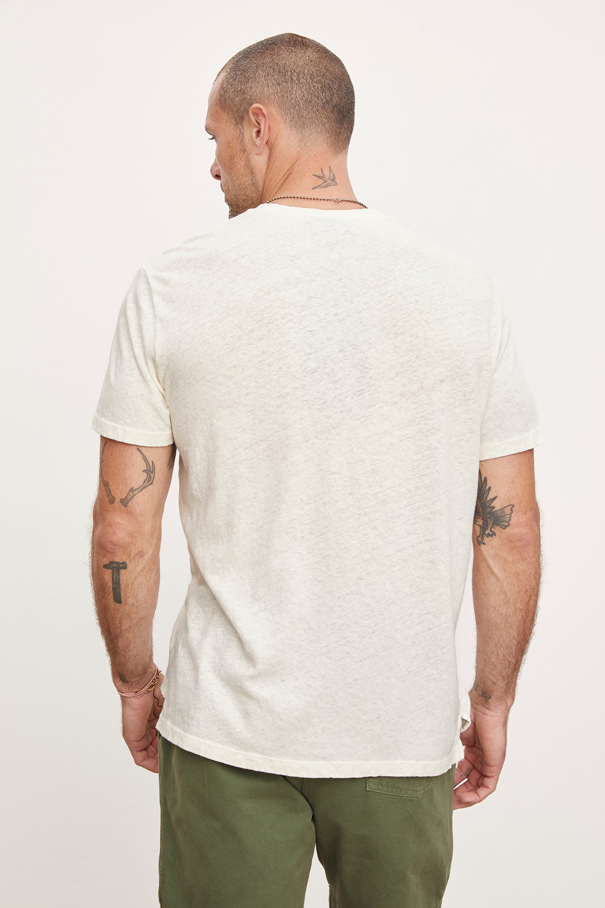 Man standing with back to the camera, showcasing various tattoos on his arms and neck, wearing a white Velvet by Graham & Spencer henley style t-shirt and green trousers.-36753589207233