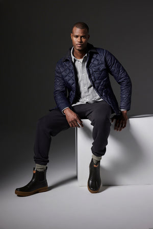 A person wearing KENJI SWEATPANTs from Velvet by Graham & Spencer, along with a quilted navy jacket, striped shirt, and boots, sits on a white block against a dark background, showcasing a vintage look.