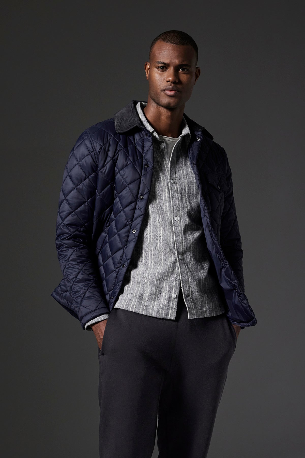 A man wearing the LOU JACKET, a dark quilted puffer jacket with a corduroy collar by Velvet by Graham & Spencer, over a light-colored shirt and dark pants stands against a dark background, with one hand in his pocket.-37888568557761