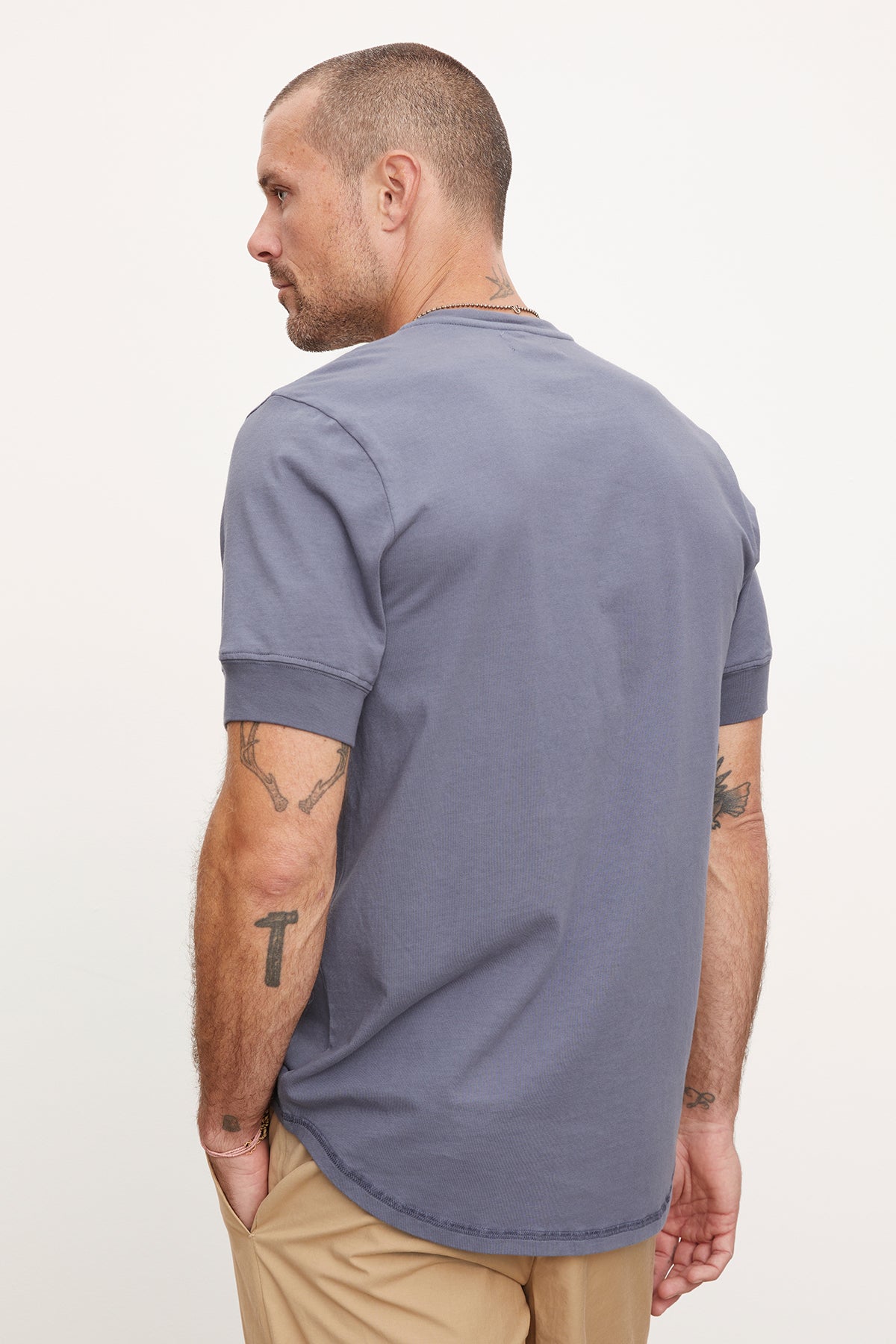   A man in a Velvet by Graham & Spencer DEON Henley t-shirt with scooped hem and beige pants, viewed from the side as he looks to the left, displaying tattoos on his arms. 