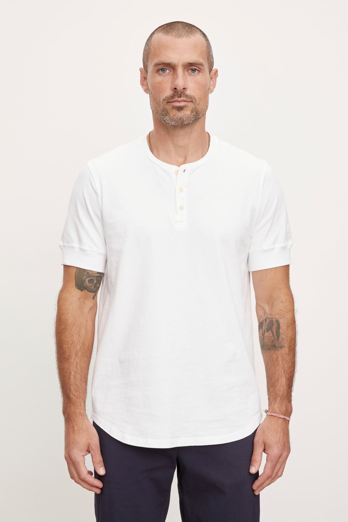   A man with a short haircut and tattoos on his arms stands against a plain background, wearing a white cotton knit DEON HENLEY by Velvet by Graham & Spencer with scooped hem details and dark pants. 