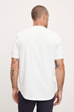 A man viewed from behind wearing a plain white short-sleeved Velvet by Graham & Spencer DEON HENLEY t-shirt and navy trousers, with visible tattoos on both arms.