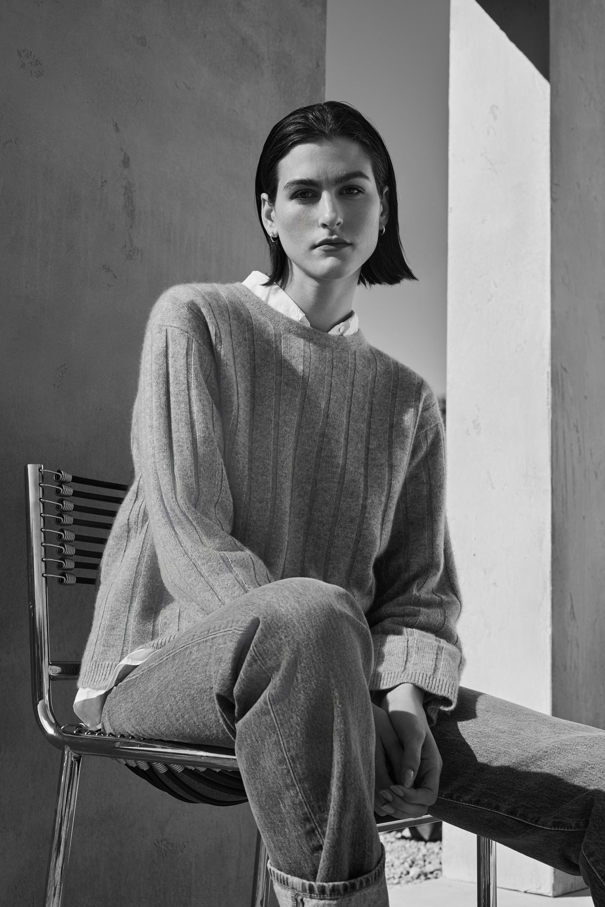   A person with short hair sits on a chair outdoors, wearing the FILLMORE CASHMERE SWEATER by Velvet by Jenny Graham and jeans. The image is in black and white. 