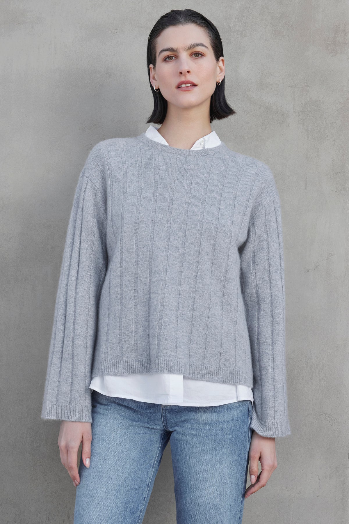   A person with dark, short hair, wearing a light gray FILLMORE CASHMERE SWEATER by Velvet by Jenny Graham layered over a white shirt and blue jeans, stands against a plain gray wall. 