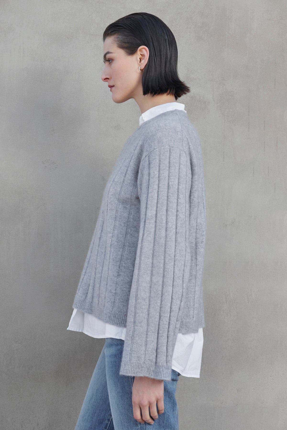 Side profile of a person wearing a grey FILLMORE CASHMERE SWEATER by Velvet by Jenny Graham over a white shirt and blue jeans, standing against a plain grey background.-37659908210881