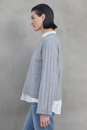 Side profile of a person wearing a grey FILLMORE CASHMERE SWEATER by Velvet by Jenny Graham over a white shirt and blue jeans, standing against a plain grey background.