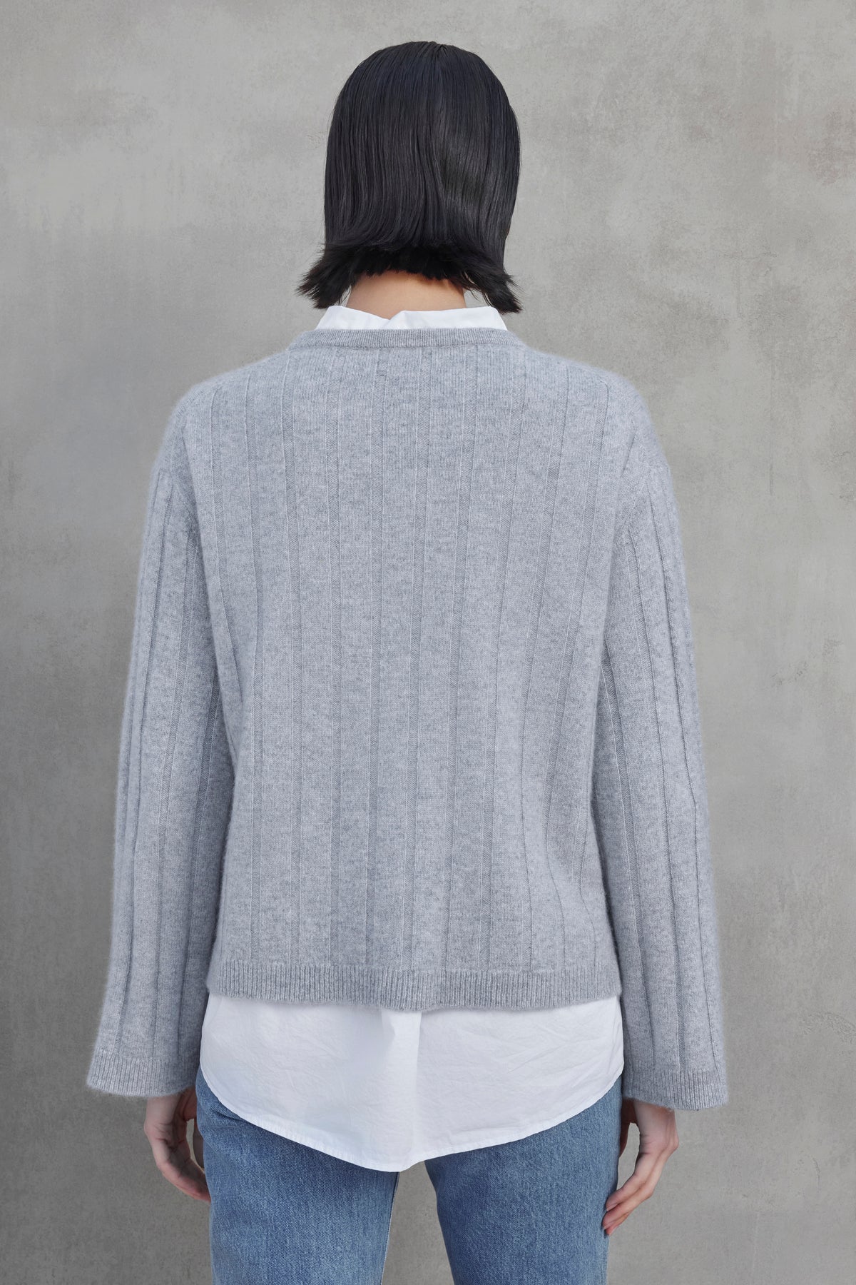   Person with short dark hair wearing the FILLMORE CASHMERE SWEATER by Velvet by Jenny Graham over a white shirt and blue jeans stands facing away against a gray background. 