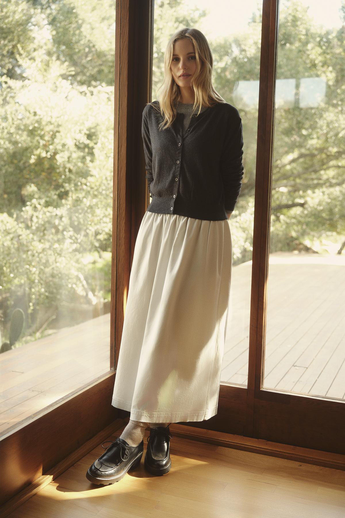   A woman stands in front of a glass door, wearing a black cardigan and the SOPHIA SKIRT made of stretch cotton twill by Velvet by Graham & Spencer, with trees and sunlight visible in the background. 