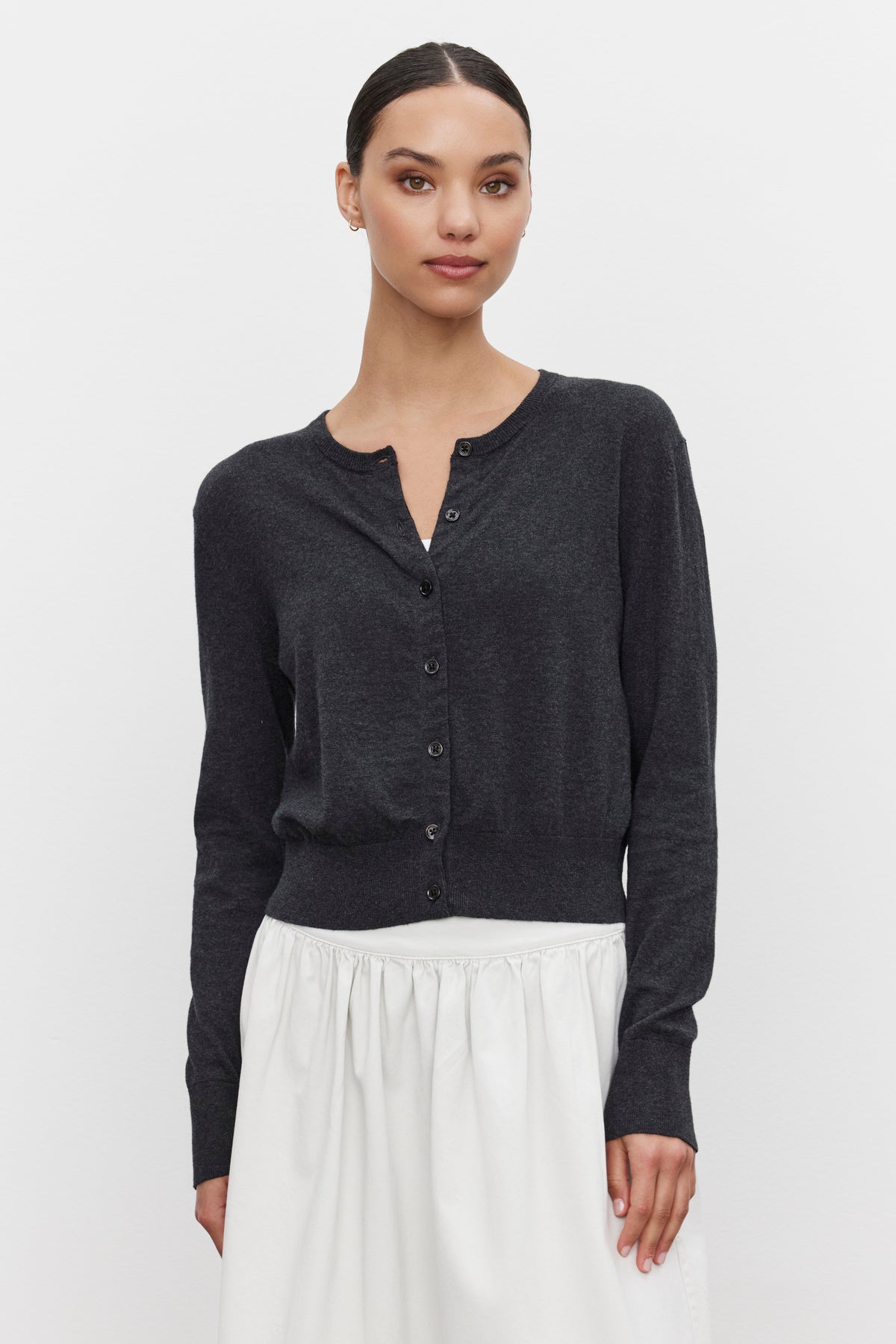   A woman stands against a white background, wearing the VIOLETTE CARDIGAN by Velvet by Graham & Spencer in dark gray lux cotton cashmere, paired with a white skirt, exuding a lightweight feel. 