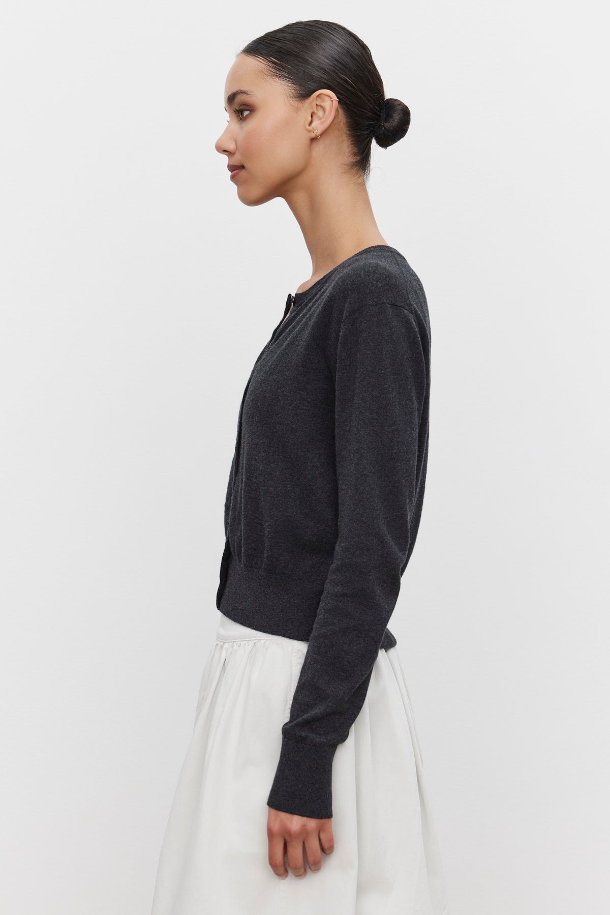   A woman with dark hair tied in a bun is standing in profile, wearing the VIOLETTE CARDIGAN by Velvet by Graham & Spencer—a charcoal gray cropped fit sweater—and a white skirt, against a plain background. 