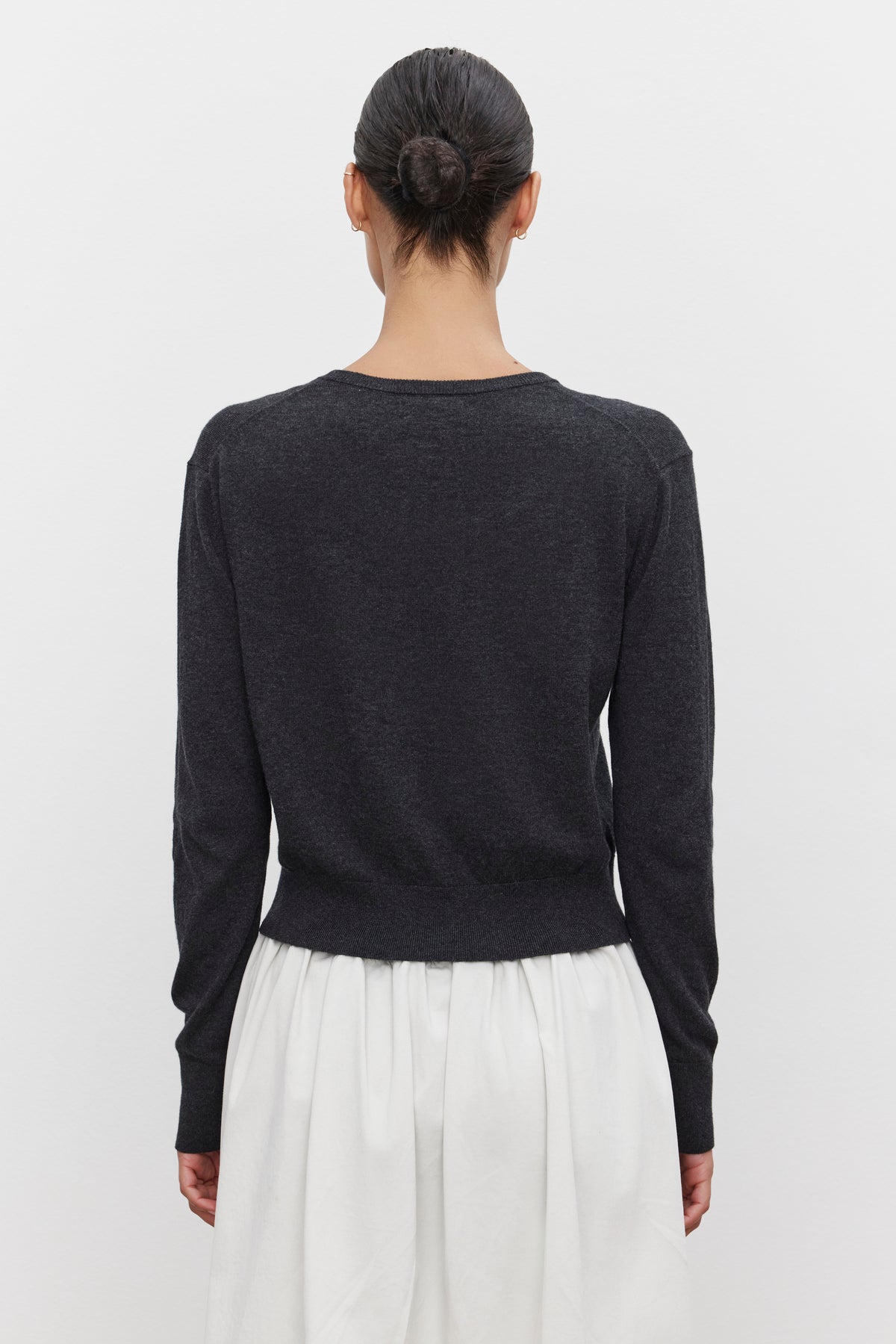   A person with dark hair in a bun stands facing away, wearing a long-sleeve black top and a white skirt, topped with the lightweight VIOLETTE CARDIGAN from Velvet by Graham & Spencer. 