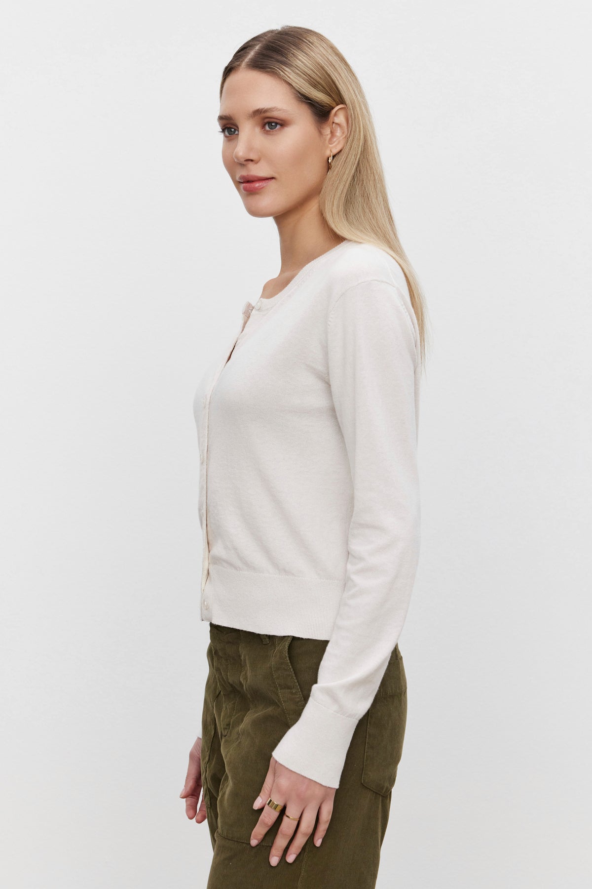   A person with long blonde hair is standing sideways, wearing the VIOLETTE CARDIGAN from Velvet by Graham & Spencer, which has a lightweight feel, paired with olive green pants. 