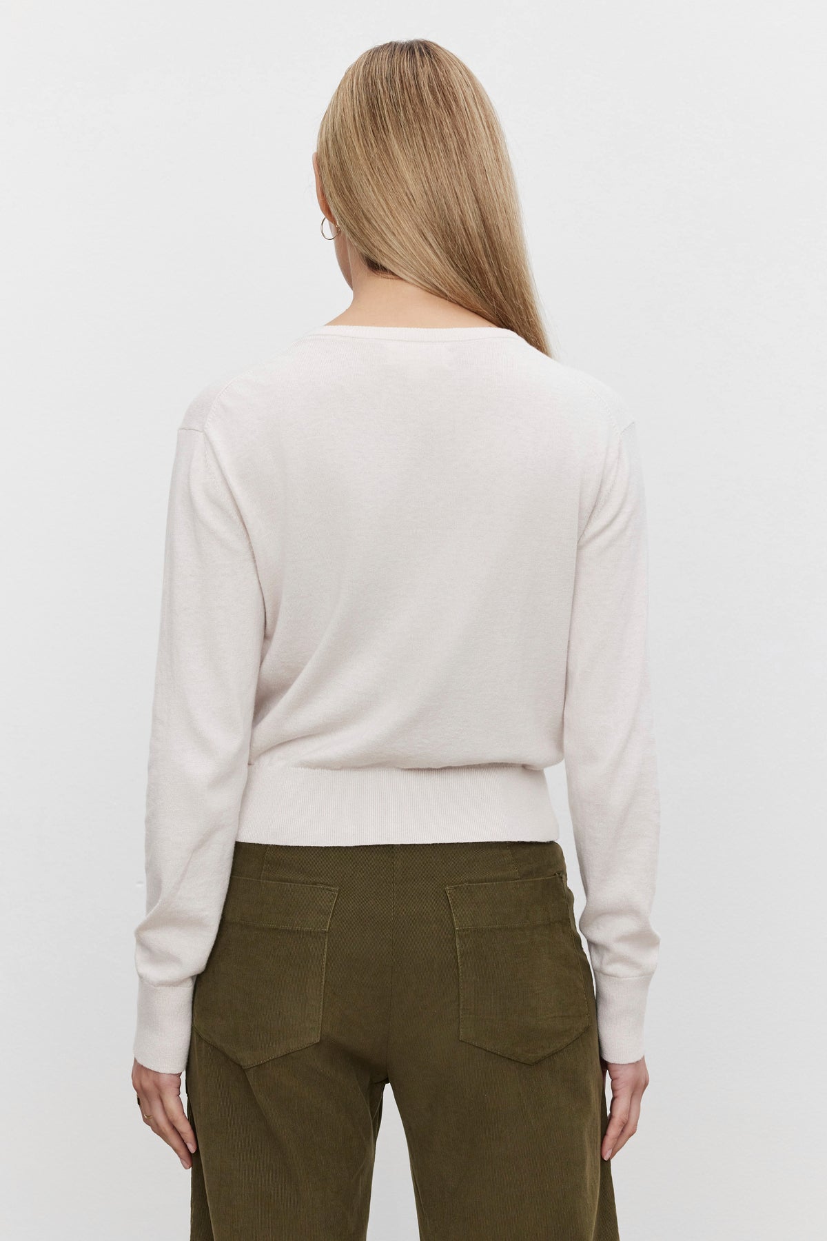   A person with long, blonde hair is shown from the back, wearing the VIOLETTE CARDIGAN by Velvet by Graham & Spencer in a light-colored, cropped fit and olive green pants, exuding a lightweight feel. 