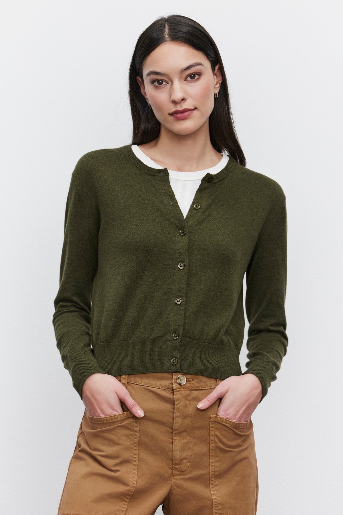 A woman with long dark hair, wearing the Velvet by Graham & Spencer Violette Cardigan in green, which features a lux cotton cashmere blend and a cropped fit over a white shirt, poses with her hands in the pockets of her tan pants against a plain white background.-37676750307521