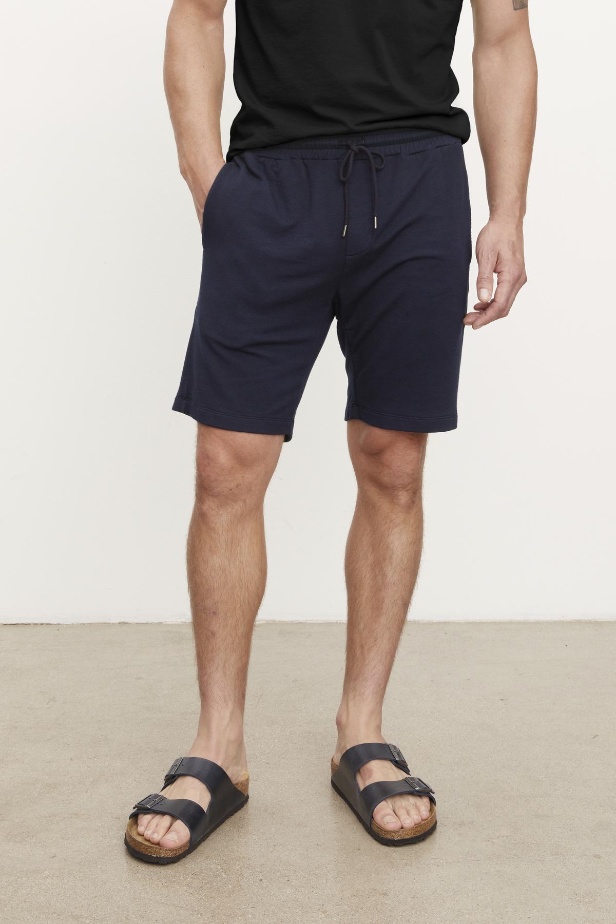 A person in an ATLAS SHORT by Velvet by Graham & Spencer and a black T-shirt stands on a concrete floor, ready for workouts, wearing sandals.-38457201721537