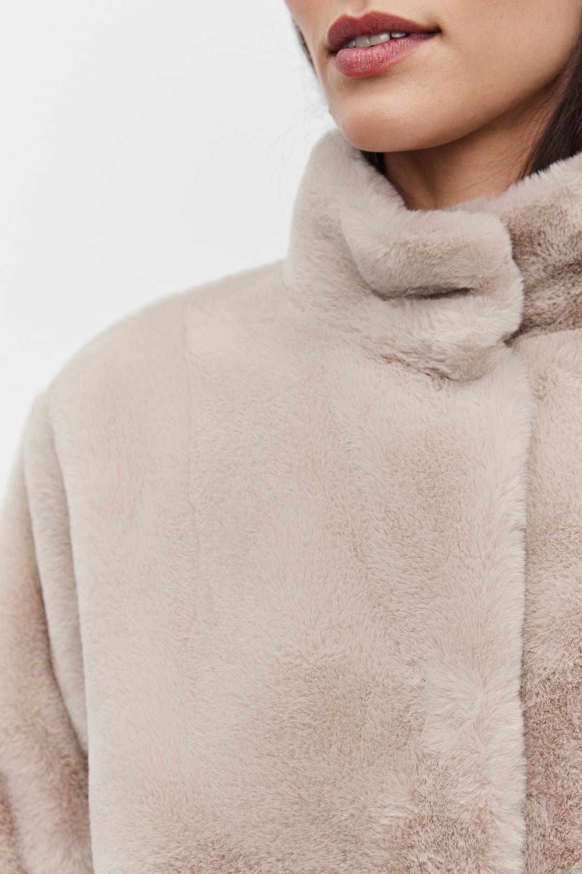   Close-up of a person wearing the SHEENA LUXE FAUX FUR JACKET by Velvet by Graham & Spencer, showcasing its plush, mock neck collar in beige. Only the lower half of their face is visible against a plain background, emphasizing the jacket's luxurious texture. 