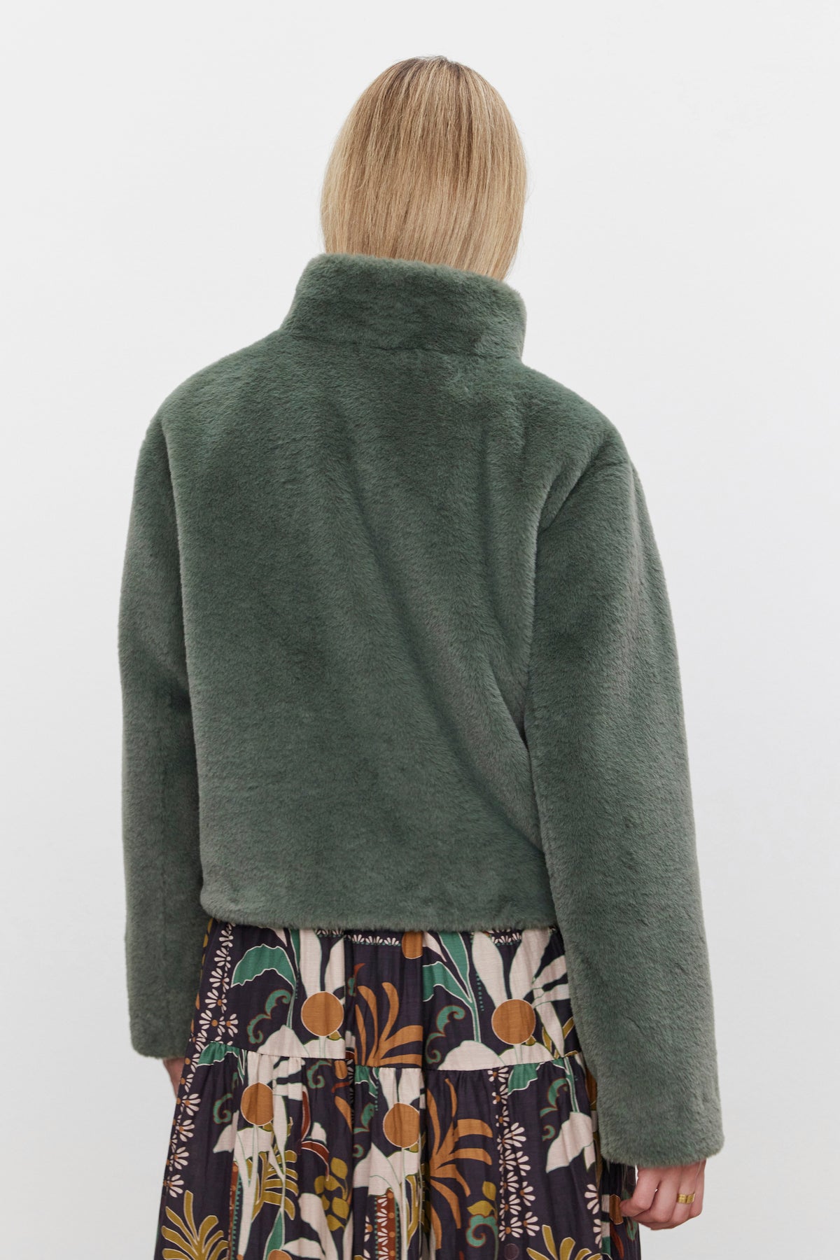   A person wearing the SHEENA LUXE FAUX FUR JACKET by Velvet by Graham & Spencer, featuring a green faux fur design and a mock neck collar, paired with a patterned skirt, is facing away from the camera against a plain background. 