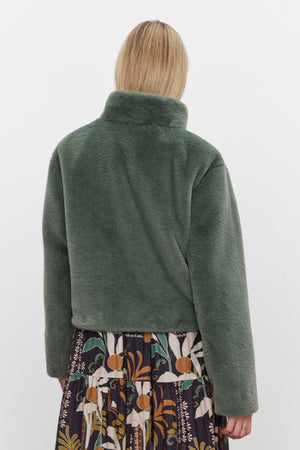 A person wearing the SHEENA LUXE FAUX FUR JACKET by Velvet by Graham & Spencer, featuring a green faux fur design and a mock neck collar, paired with a patterned skirt, is facing away from the camera against a plain background.