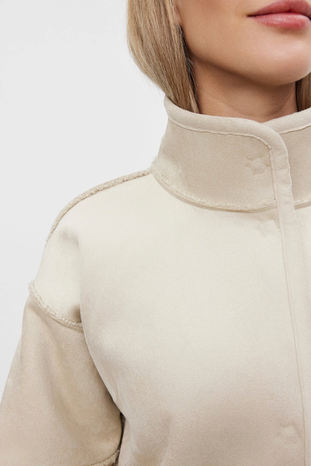 Close-up of a person wearing the CARA LUXE SHERPA REVERSIBLE JACKET in light beige, featuring a high collar and prominent seams. The jacket by Velvet by Graham & Spencer, made with faux suede material, boasts a soft texture against the plain white background.-37893996871873