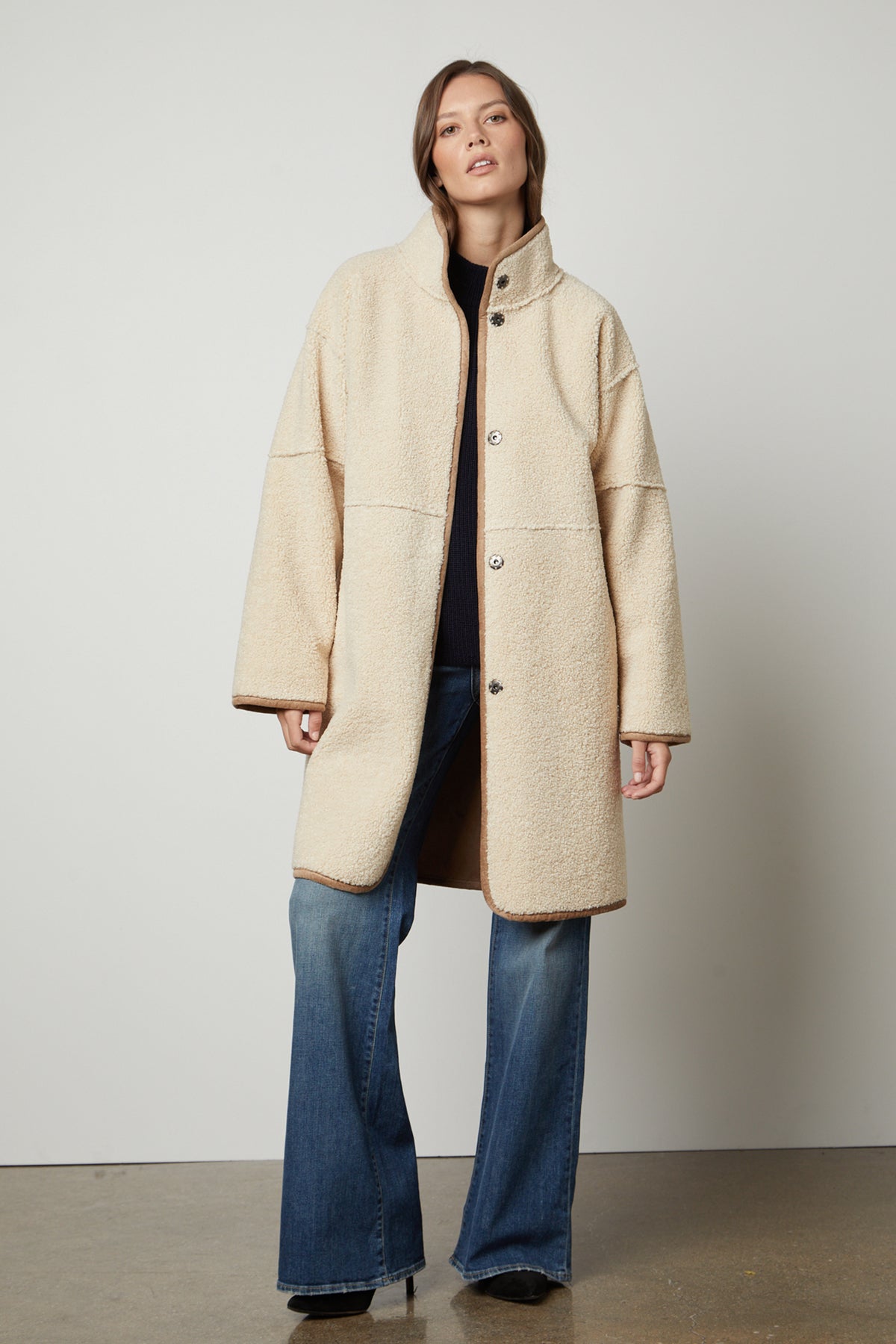   A person wearing a long beige CARA LUXE SHERPA REVERSIBLE JACKET by Velvet by Graham & Spencer and blue jeans stands against a plain background. 