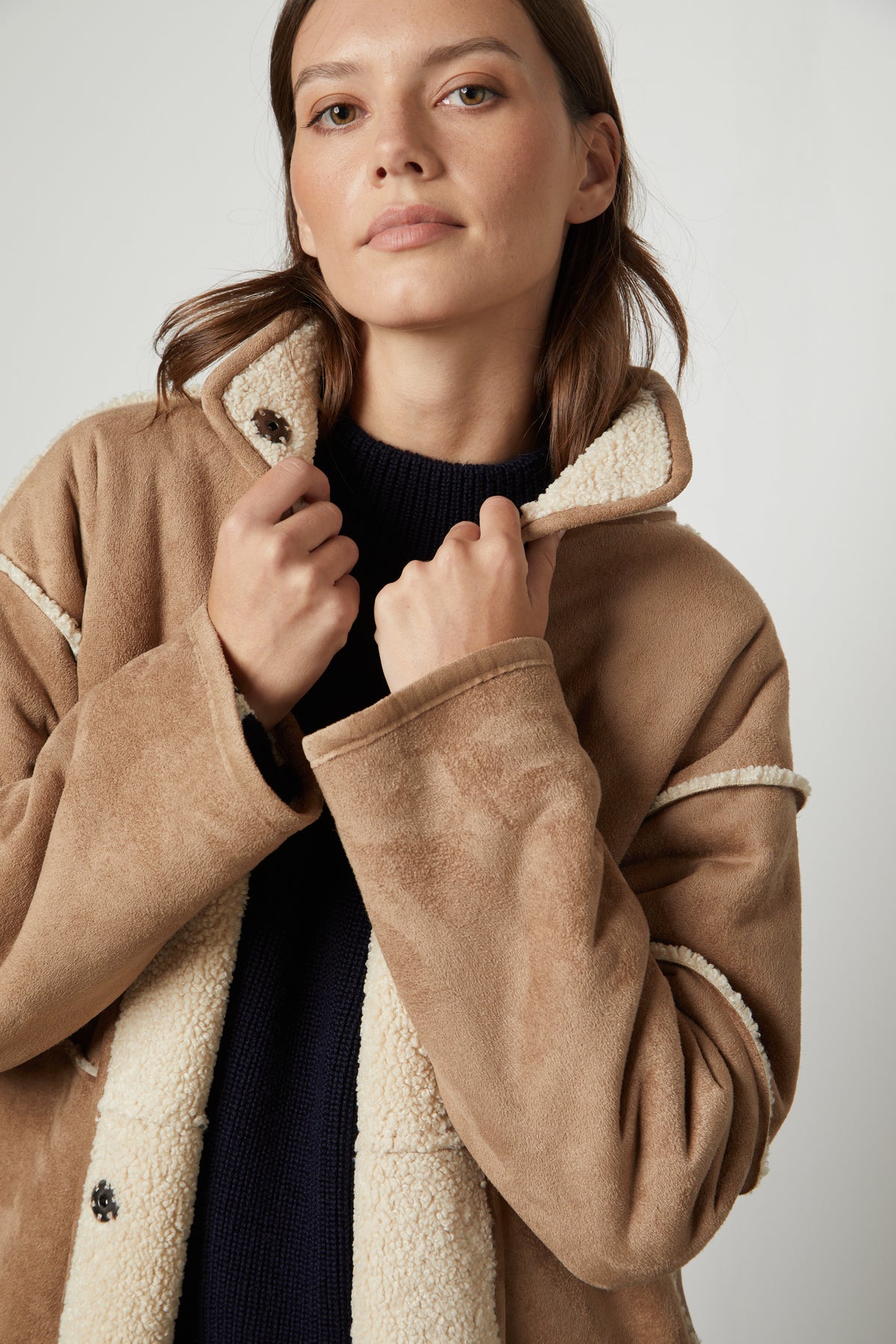   A person wearing the CARA LUXE SHERPA REVERSIBLE JACKET by Velvet by Graham & Spencer, with a black sweater underneath, looks directly at the camera. 