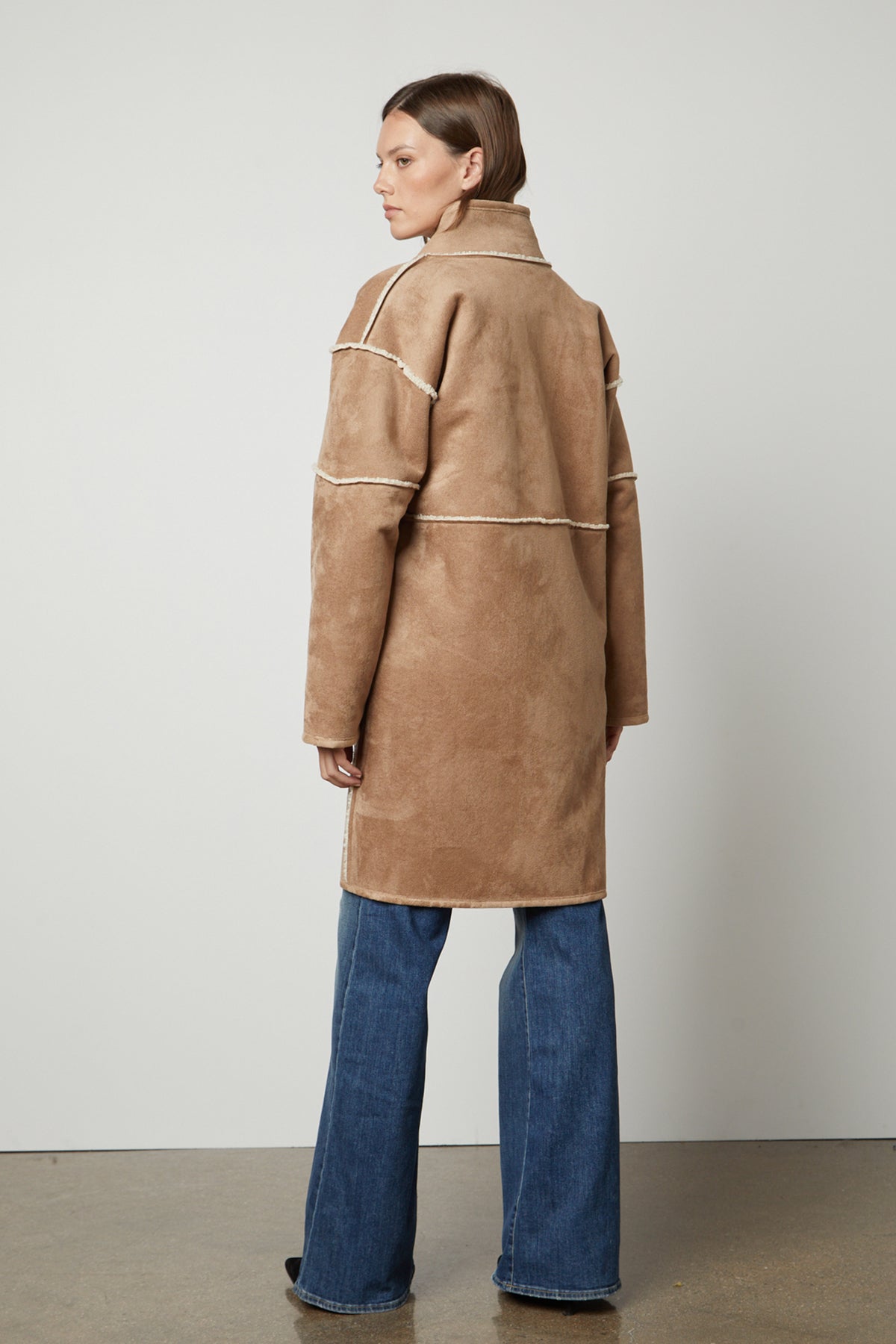 A person wearing a long tan CARA LUXE SHERPA REVERSIBLE JACKET by Velvet by Graham & Spencer and blue jeans stands facing away against a plain background.-26897724047553