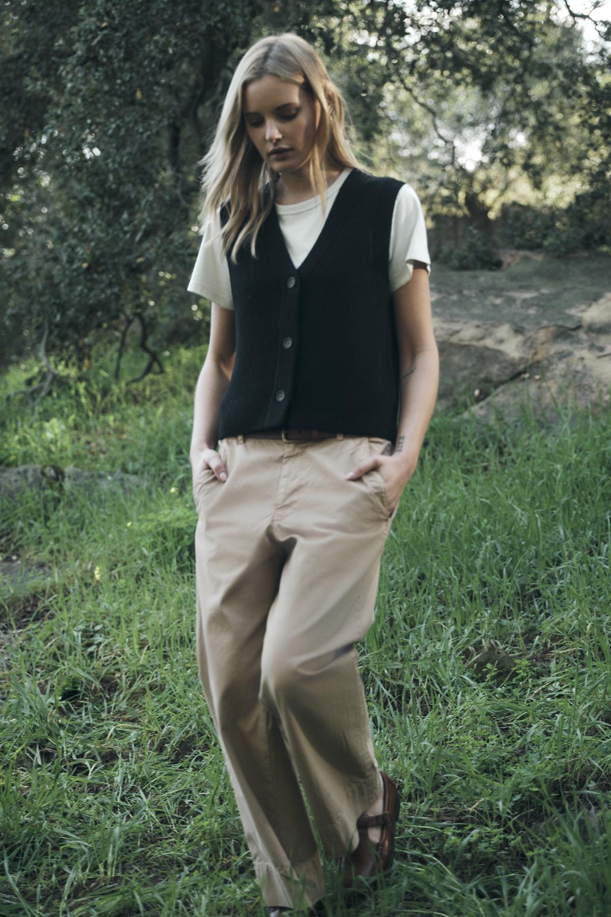 A person stands outdoors in casual attire, wearing a white V-neck t-shirt, the LU SWEATER VEST by Velvet by Graham & Spencer in black wool blend, and beige pants, with hands in pockets. Green grass and trees are in the background.-37676225102017