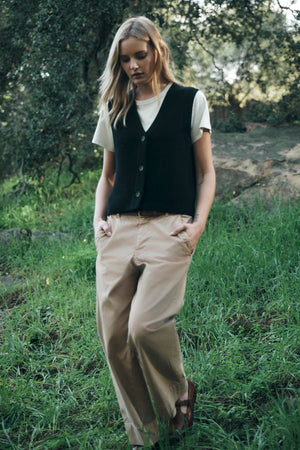 A person stands outdoors in casual attire, wearing a white V-neck t-shirt, the LU SWEATER VEST by Velvet by Graham & Spencer in black wool blend, and beige pants, with hands in pockets. Green grass and trees are in the background.