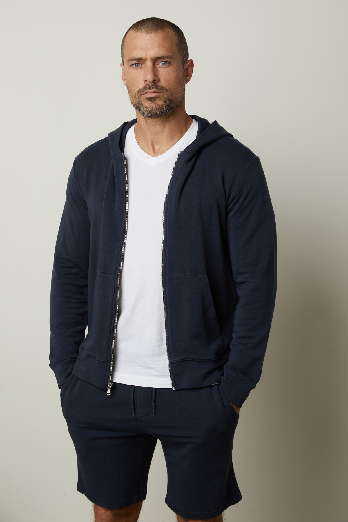   A man with a shaved head and full beard is wearing a RODAN HOODIE by Velvet by Graham & Spencer, a luxe fleece navy blue zip-up hoodie, over a white t-shirt and matching navy blue shorts, standing against a plain background. 