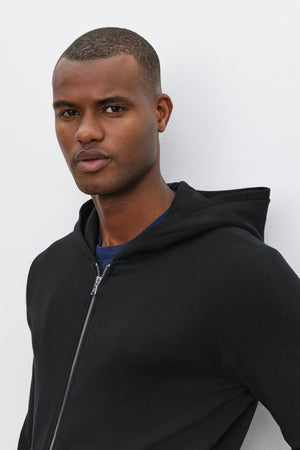 A person wearing a RODAN HOODIE by Velvet by Graham & Spencer, which features a black zip-up design with a cozy brushed interior, along with a dark shirt, stands against a white background.