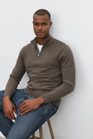 A person wearing the FLETCHER QUARTER-ZIP SWEATER from Velvet by Graham & Spencer in brown over a white shirt and blue jeans sits on a stool against a plain white background.