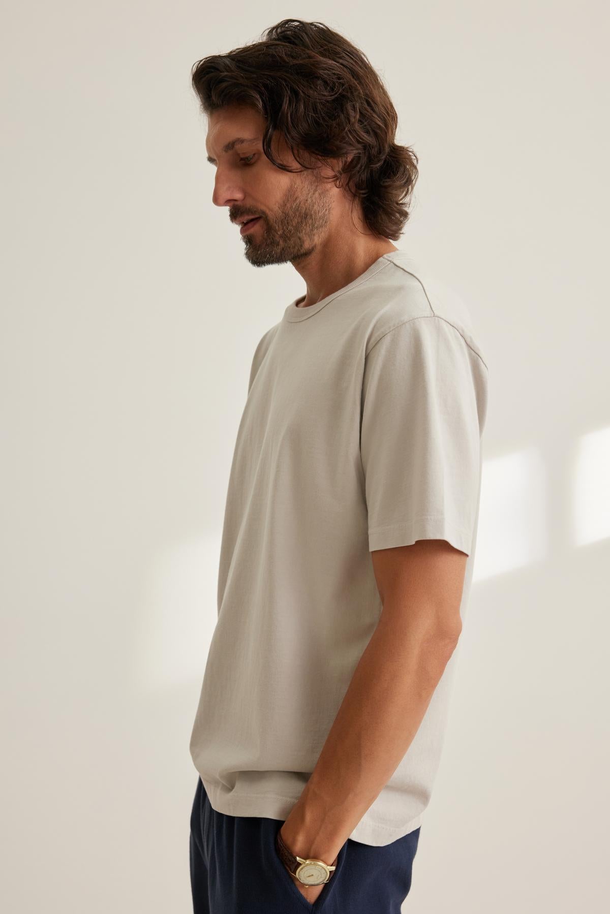   A man with shoulder-length hair and a beard stands in profile, wearing a Velvet by Graham & Spencer WELLS TEE, white in color, along with dark pants, and has his hands in his pockets. 