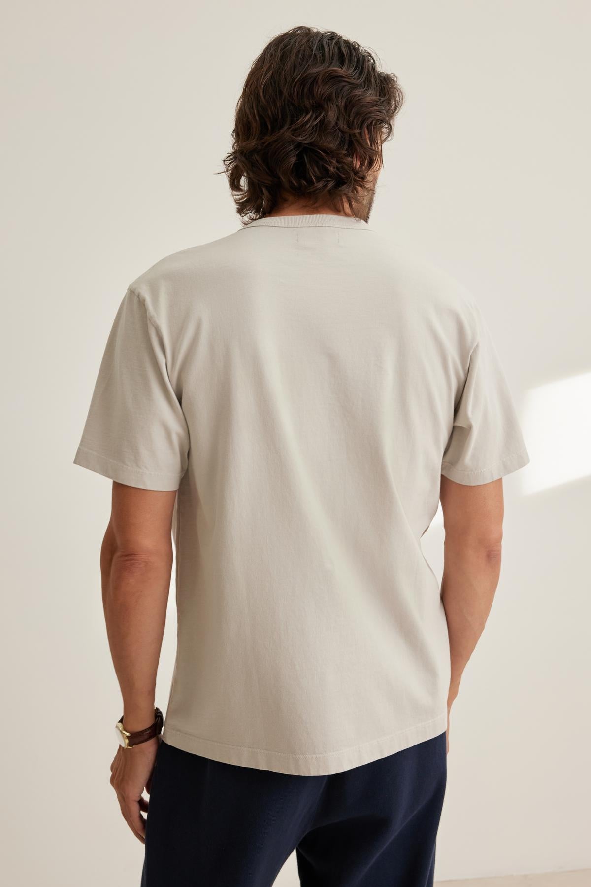   A person with wavy hair stands facing away, wearing the WELLS TEE by Velvet by Graham & Spencer in light gray, paired with dark pants against a plain background. 