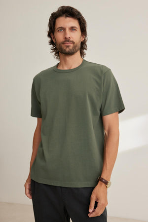 A man with dark hair and a beard wears the WELLS TEE by Velvet by Graham & Spencer, paired with black pants, standing against a plain beige background.