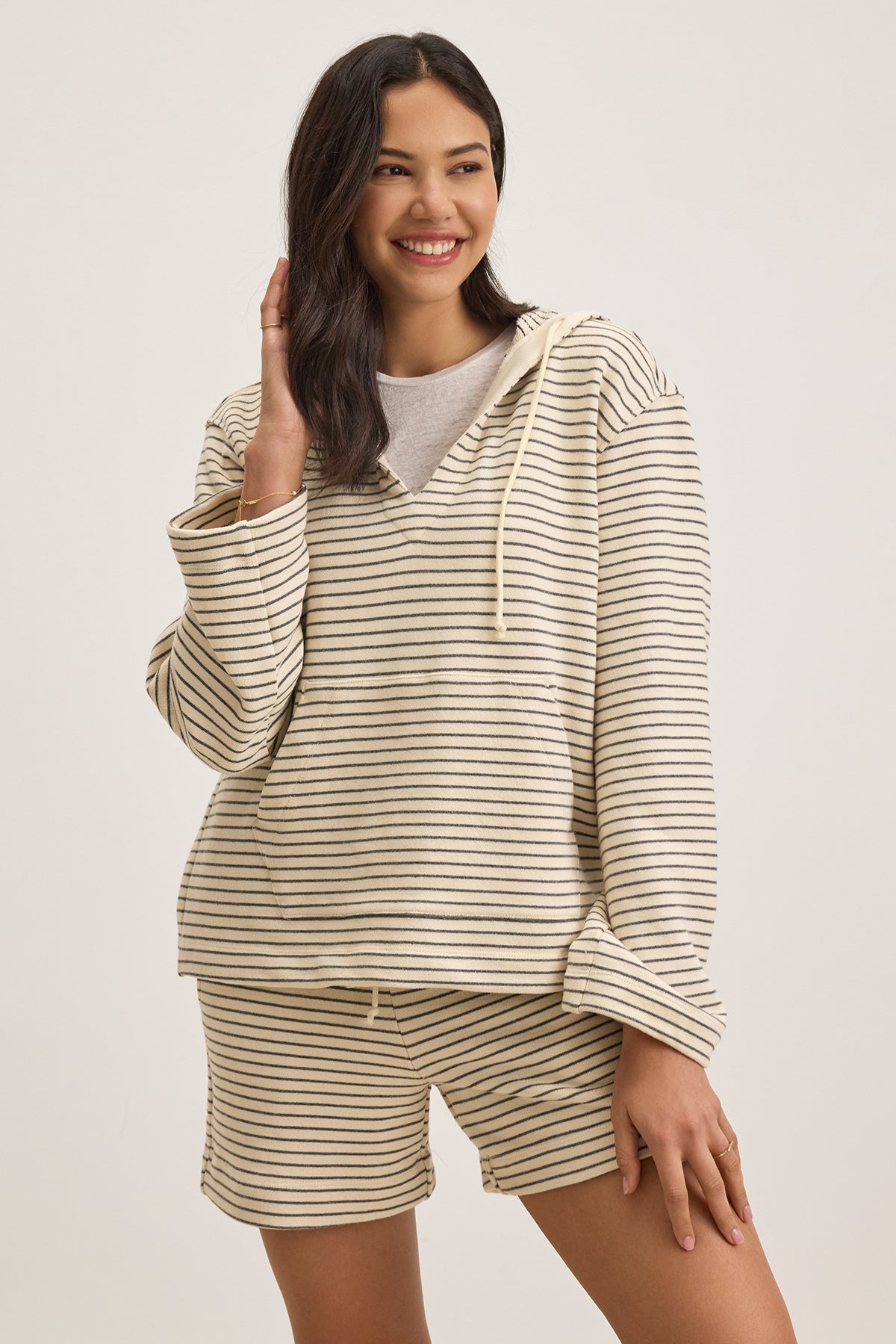 A person smiles while touching their hair, wearing the JORDYN HOODIE by Velvet by Graham & Spencer—a beige and black striped mid-weight French terry loungewear essential—paired with shorts against a plain background, effortlessly combining comfort and style.-38662768984257