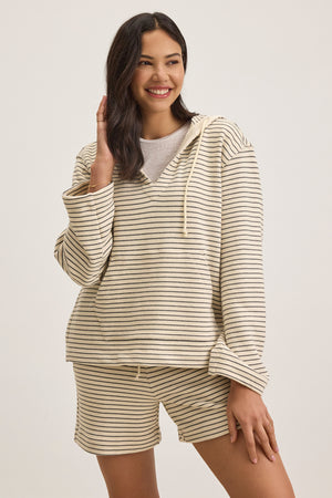 A person smiles while touching their hair, wearing the JORDYN HOODIE by Velvet by Graham & Spencer—a beige and black striped mid-weight French terry loungewear essential—paired with shorts against a plain background, effortlessly combining comfort and style.