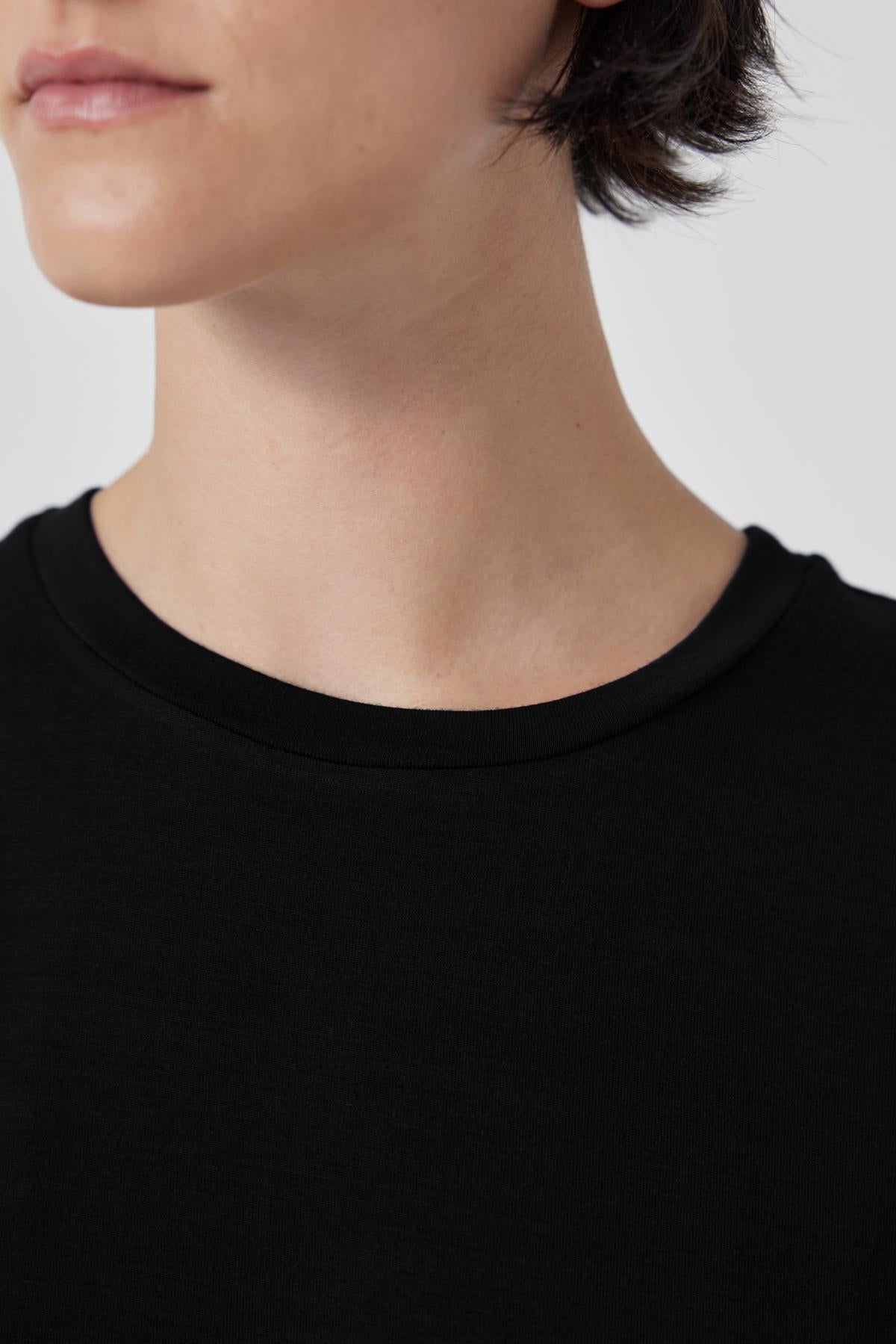 Close-up view of a person wearing a black SOLANA TEE by Velvet by Jenny Graham with short hair, partially showing their face against a plain, light background. The shirt's tailored finish highlights the soft modal jersey fabric.-36463664005313