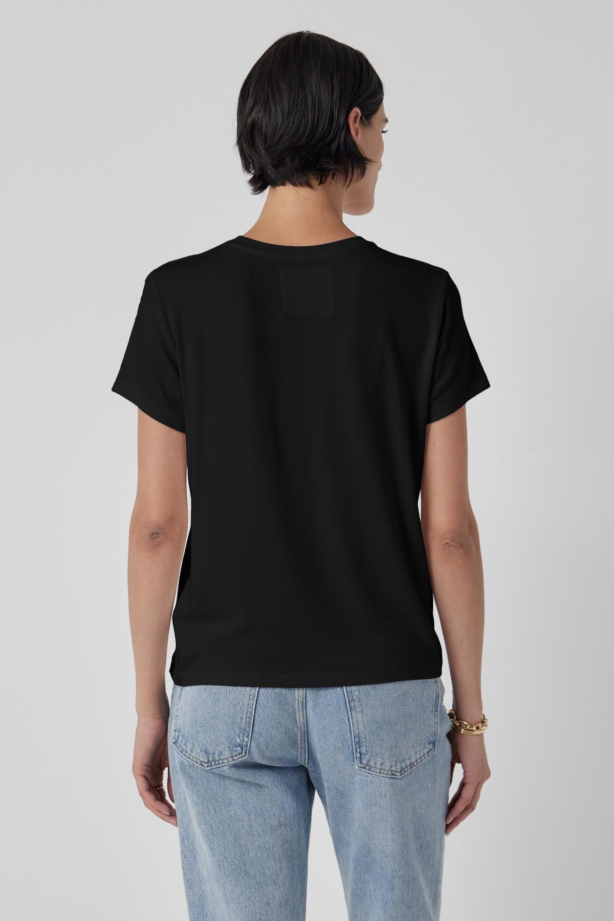   A person with short dark hair, wearing a tailored finish black SOLANA TEE by Velvet by Jenny Graham and light blue jeans, is standing with their back to the camera against a plain background. 