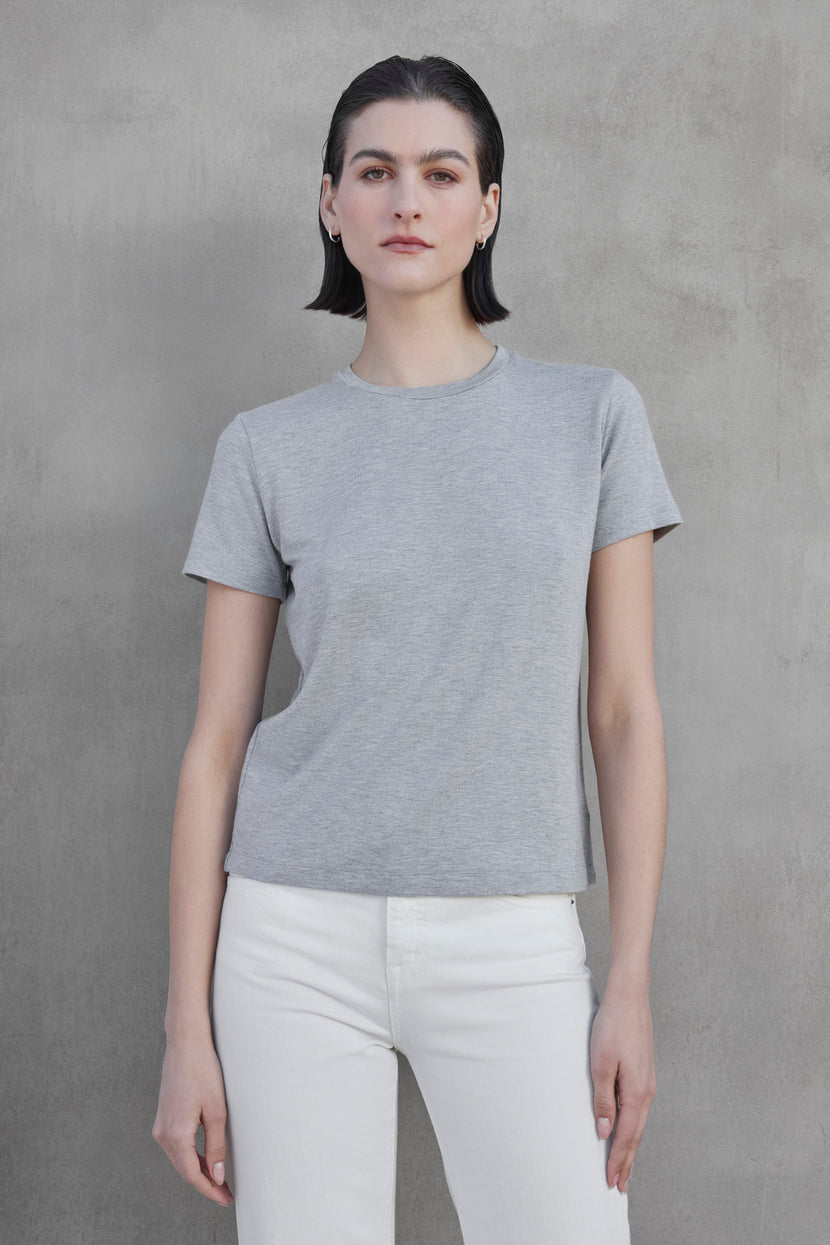 A person with short dark hair is wearing a gray SOLANA TEE by Velvet by Jenny Graham and white pants, standing against a bare, textured gray wall.