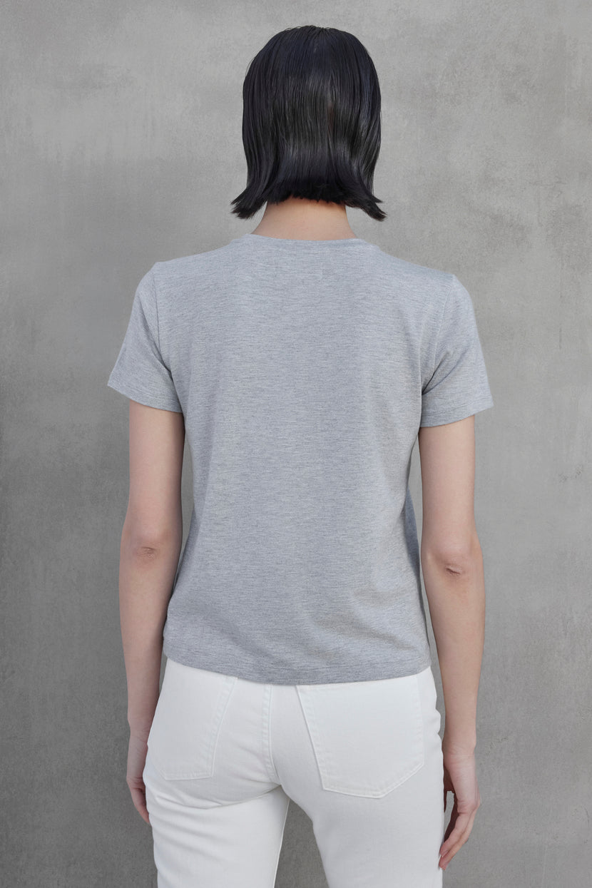 Person wearing a gray SOLANA TEE from Velvet by Jenny Graham and white pants, facing away, against a grey wall.