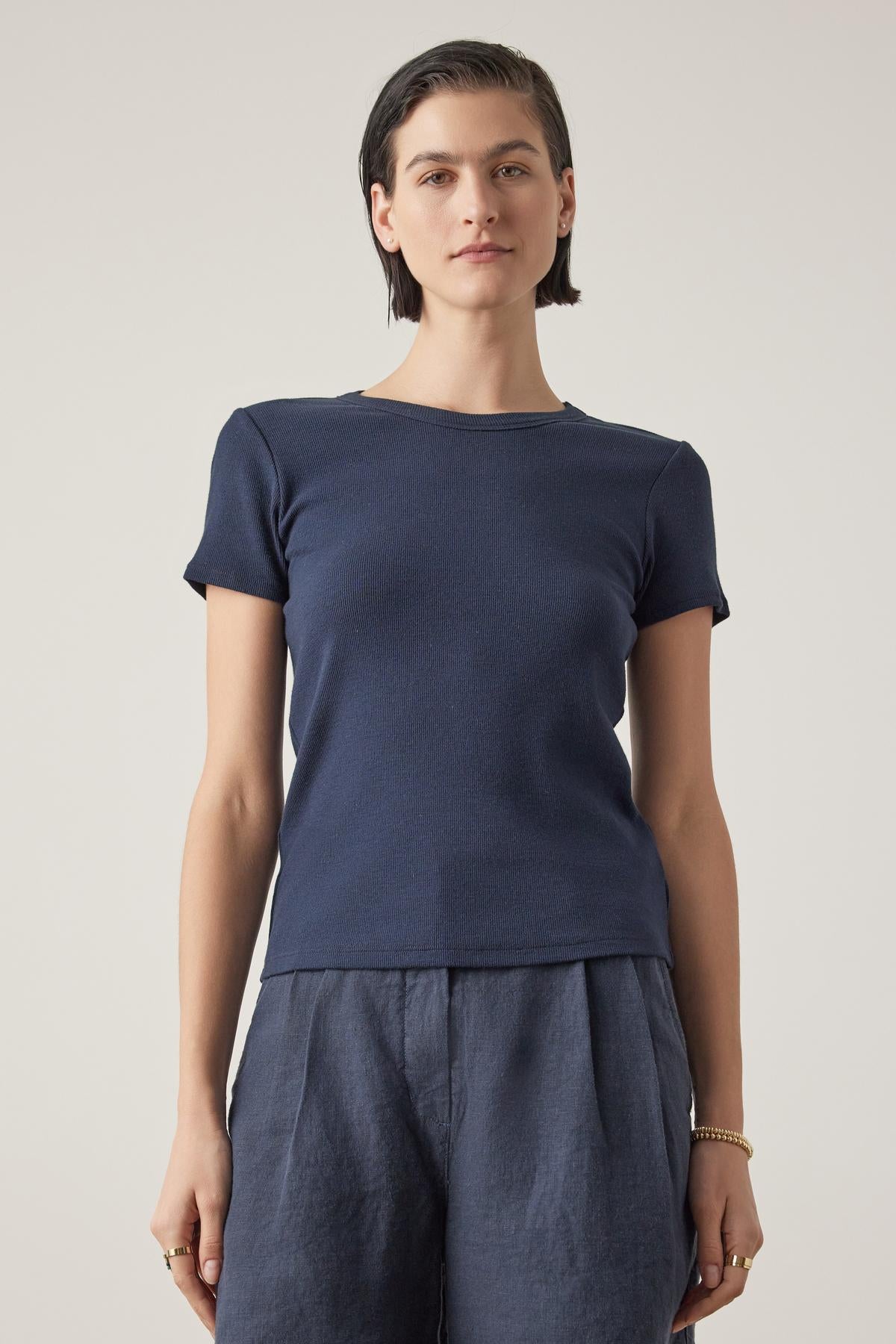   A woman with short hair, wearing a Velvet by Jenny Graham dark blue Bedford Tee with a crew neckline and gray pants, stands against a light background. 