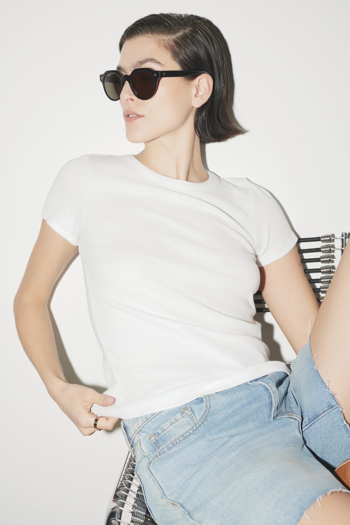 A woman in sunglasses, a white Velvet by Jenny Graham BEDFORD TEE with a crew neckline, and jeans, sitting on a metal chair against a white background, looking to the side.-36753616732353