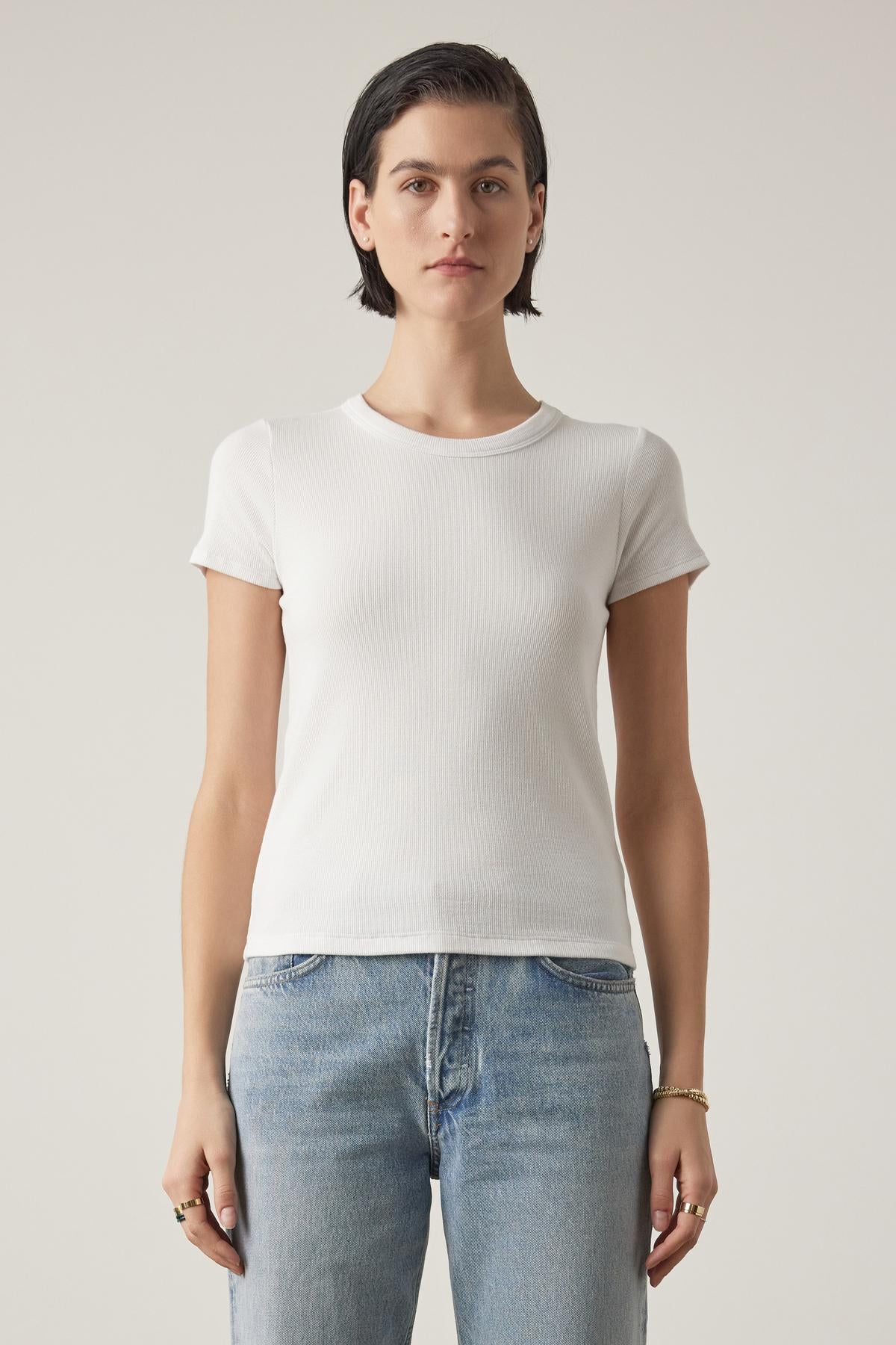   A woman with short dark hair wearing a BEDFORD TEE by Velvet by Jenny Graham in white with a crew neckline and blue jeans stands against a neutral background, facing the camera with a neutral expression. 